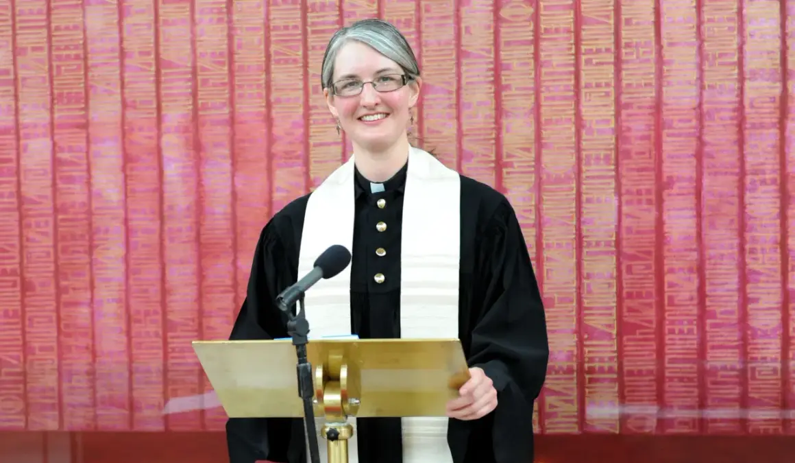 Eine Pfarrerin mit Brille und grauem Haar steht lächelnd hinter einem goldenen Pult. Sie trägt eine schwarze Robe und ein weißes Stola. In dir steckt ein guter Christ.