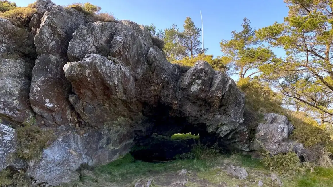 Entrada a la cova da serpe, una cueva natural entre rocas y vegetación bajo un cielo azul.