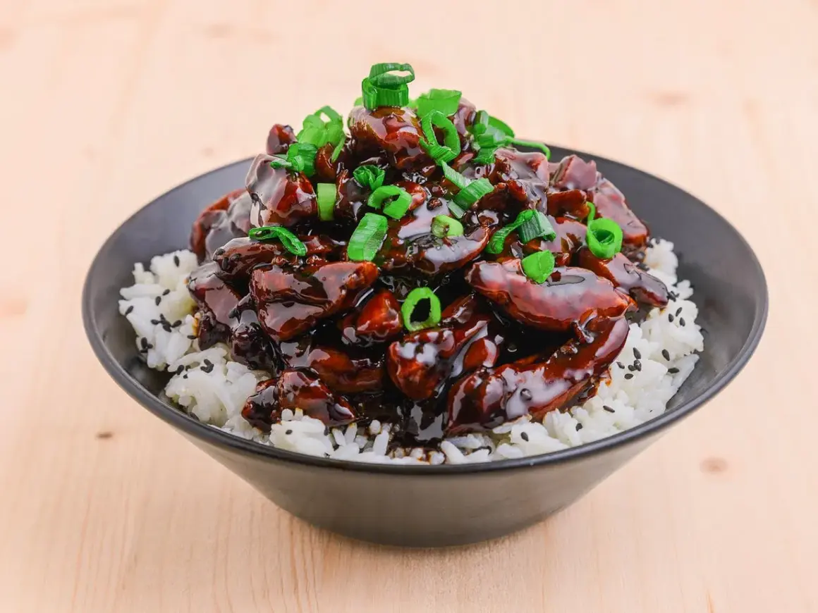 En skål med ris toppad med glansig teriyaki kyckling och strimlad salladslök.
