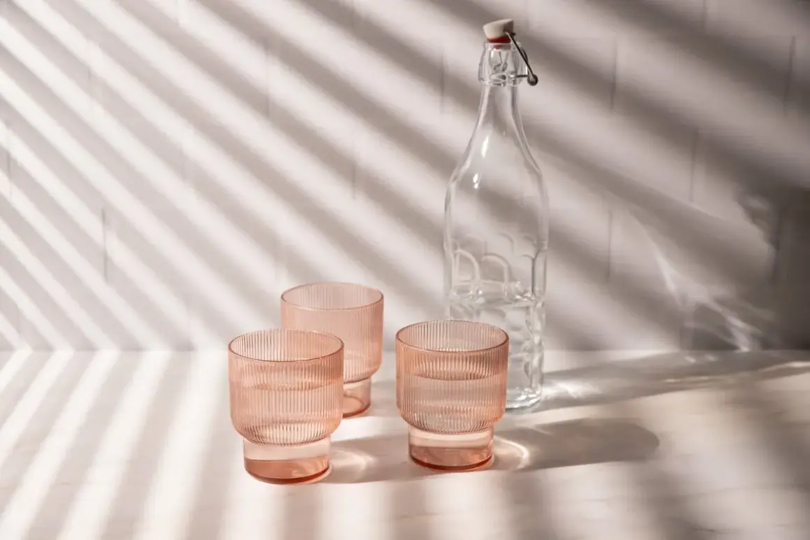 Natural product photography: three pink ribbed glasses filled with water and a clear glass bottle with a swing top, bathed in dappled light.