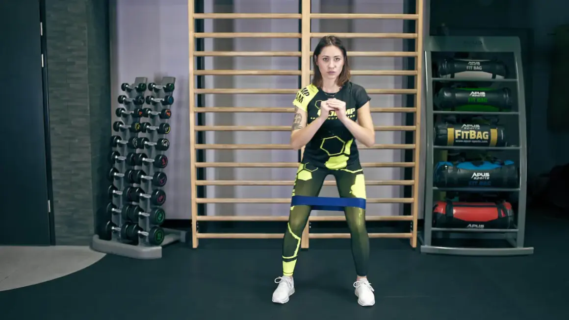 Kobieta w sportowym stroju wykonuje ćwiczenie monster walk z gumą oporową na udach.