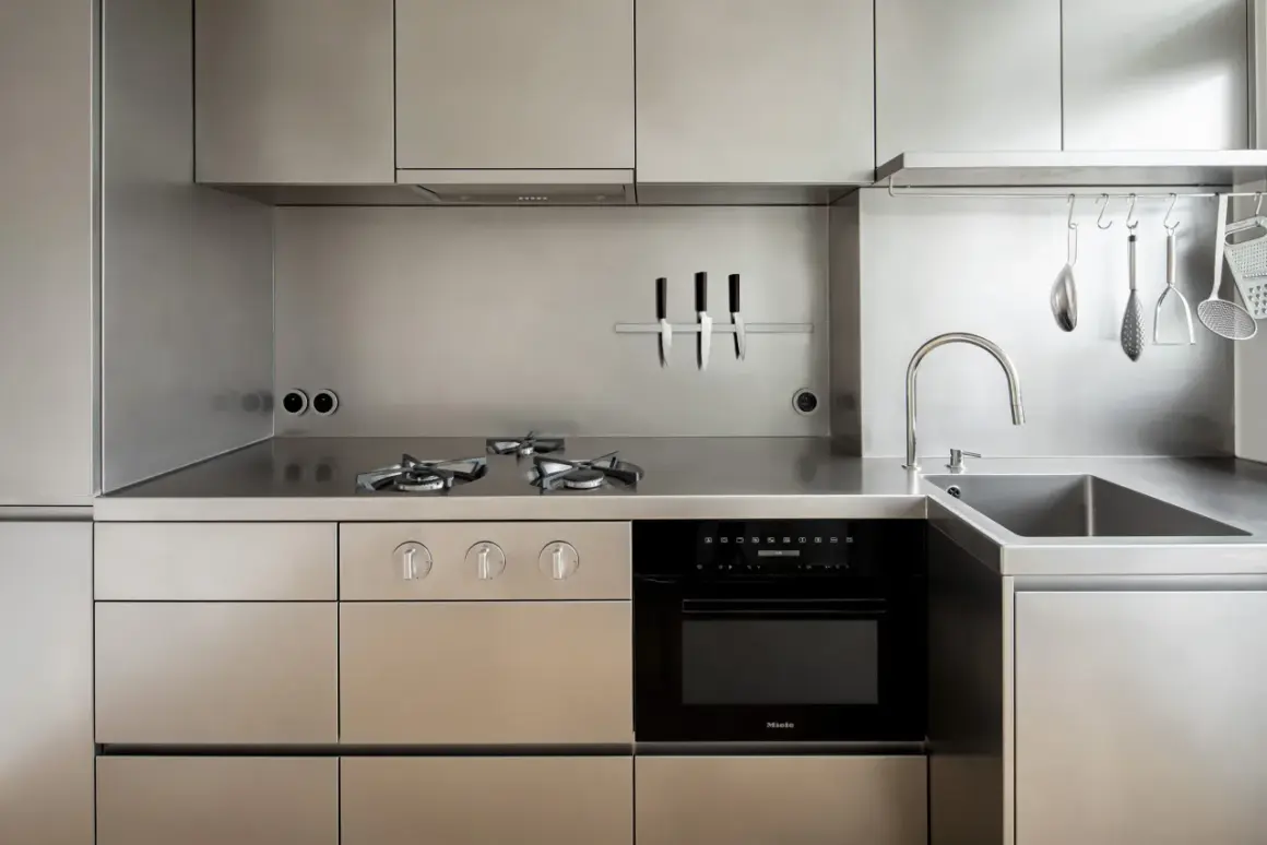 Cucina moderna con piano cottura a gas, forno, lavello e utensili appesi, tutto in lucido acciaio inox 18/10.