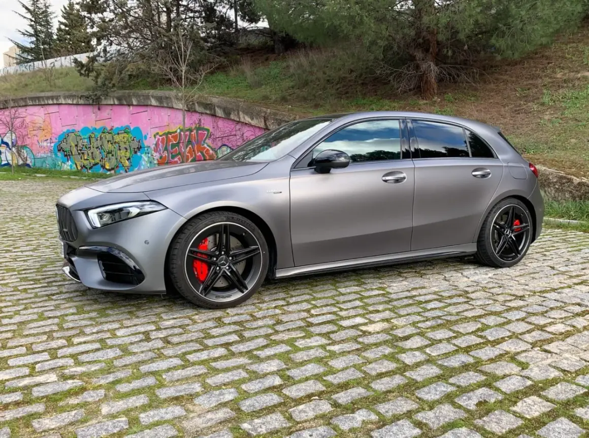 A45 AMG CV gris mate, con pinzas rojas y llantas negras, aparcado sobre adoquines frente a un muro con grafitis.