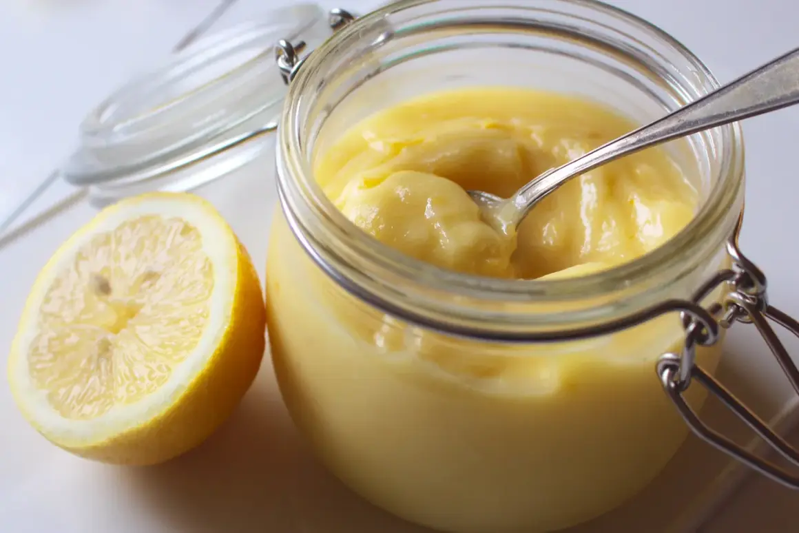 En burk med krämig lemon curd och en halv citron bredvid. En sked vilar i burken, redo att skopa upp den syrliga godsaken.