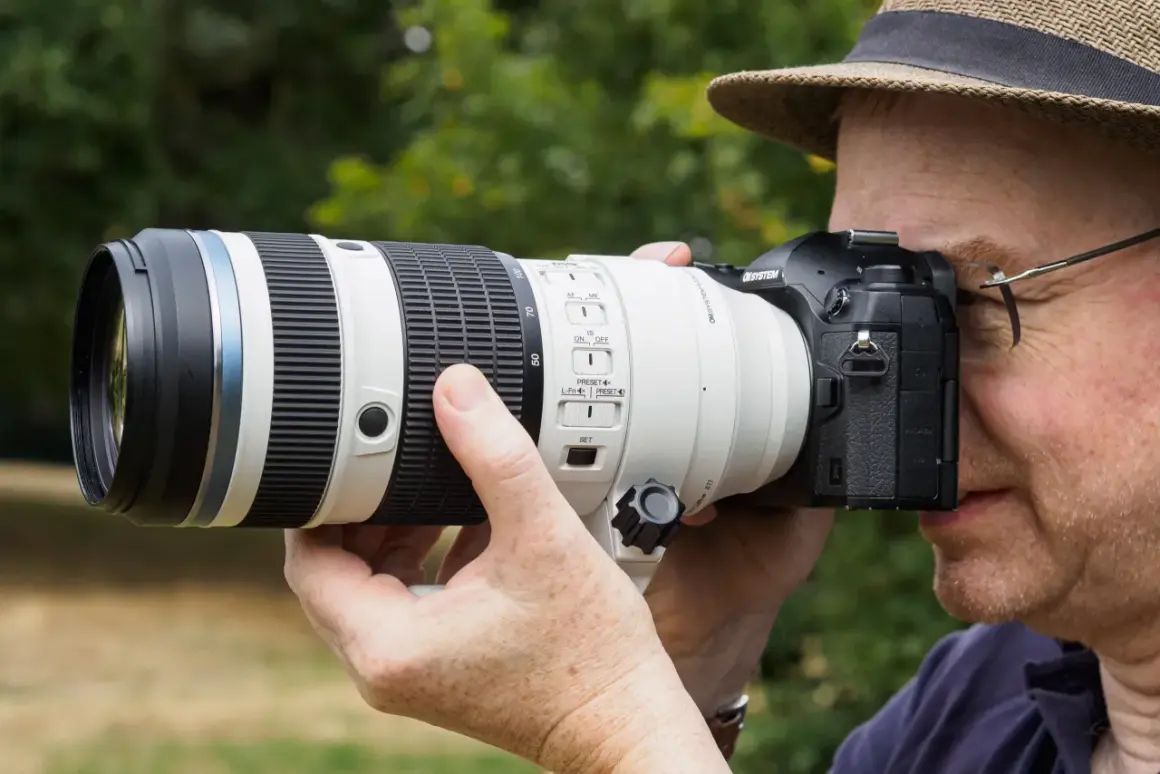 A photographer in a hat uses a large telephoto lens on a Micro Four Thirds camera, perhaps for a detailed micro 4/3 lenses review.