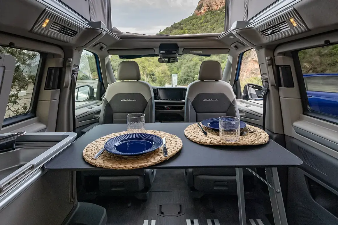 Interior de VW California listo para cenar. Mesa puesta con platos y vasos, asientos delanteros y vistas al exterior.