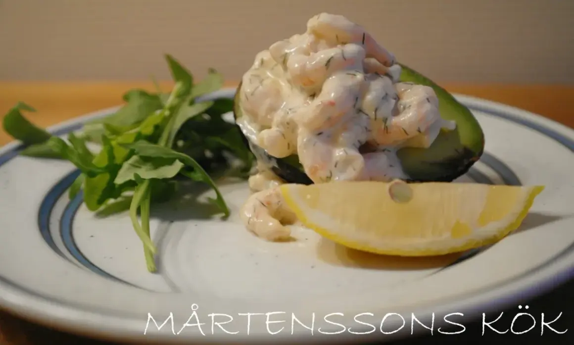 Avokadohalva fylld med räkor i en krämig sås, liknande guacamole crème fraiche, med en citronskiva och gröna blad.