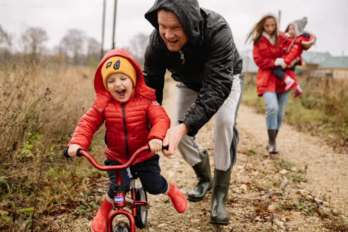 Radosne dziecko na rowerku biegowym, kiedy dziecko stoi bez podparcia, z tatą asekurującym. W tle mama z drugim dzieckiem.