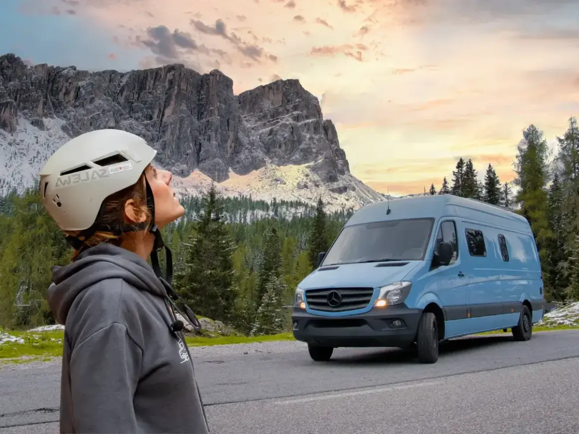 Mujer con casco mira las montañas. A su lado, un **autobús camperizado** azul espera para la aventura.