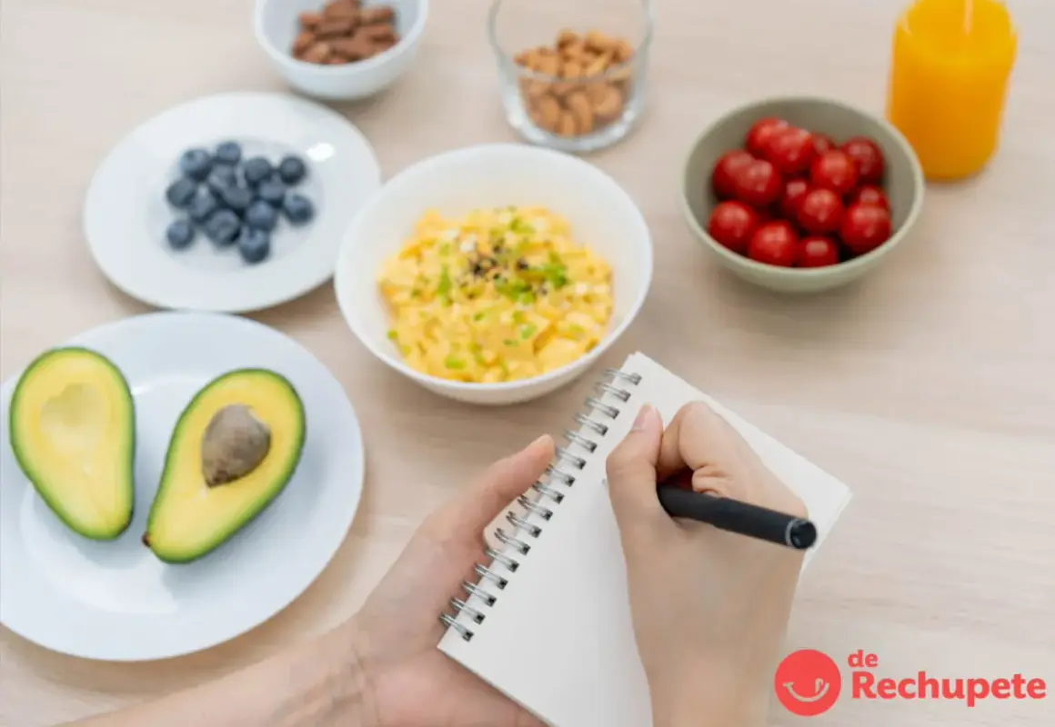 Manos escribiendo en un cuaderno, rodeadas de ingredientes para crear ejemplos de menú: aguacate, arándanos, huevos revueltos, tomates cherry, frutos secos y jugo.