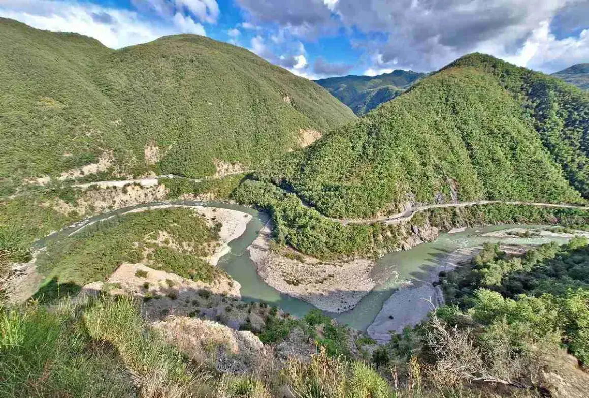 Un fiume serpeggia tra montagne verdi e scoscese, con una strada che costeggia il corso d'acqua. Il borgo di Brugnello si intravede tra la vegetazione lussureggiante.