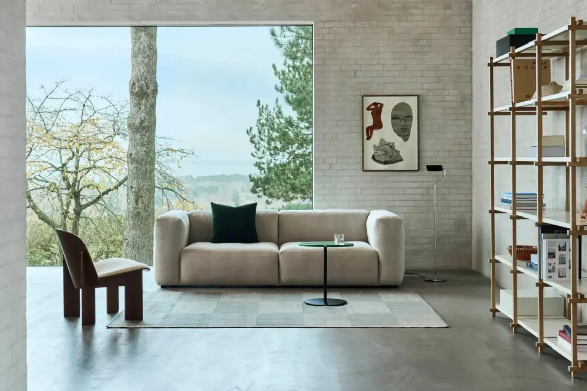Gemütliches Wohnzimmer mit einem Sofa in millennial grey, einem Holzsessel und einem Bücherregal vor einem Fenster mit Blick ins Grüne.