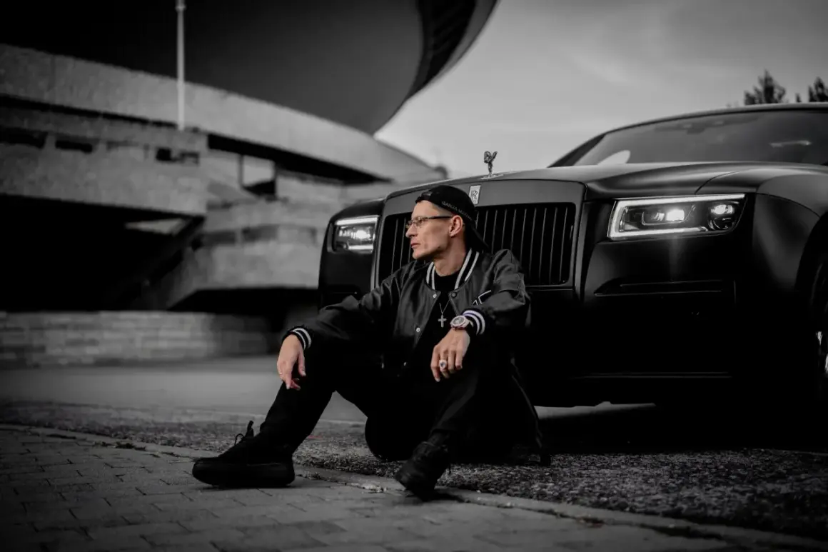 Mężczyzna w czapce i kurtce siedzi obok czarnego Rolls-Royce'a na tle betonowej budowli.