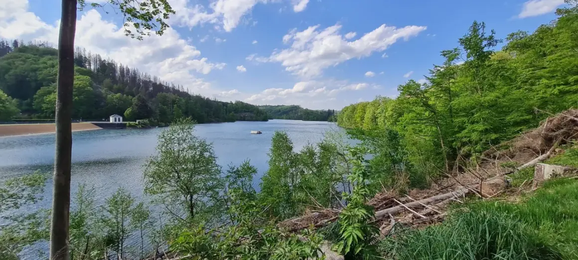 Ein ruhiger See mit einem kleinen Haus am Ufer, umgeben von grünen Wäldern. Der genkeltalsperre rundweg lädt zum Spaziergang ein.