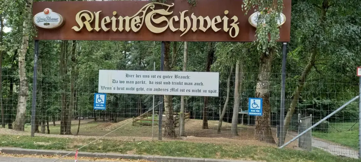 Schild "Kleine Schweiz" in Velbert. Ein Spruch lädt zum Verweilen ein. Parken, essen, trinken – perfekt für eine Wanderung.