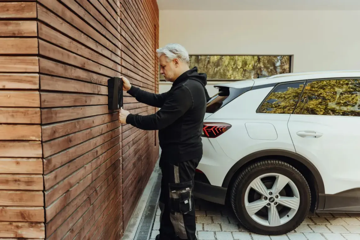 Un homme installe une borne de recharge pour voiture électrique sur un mur en bois. La question "peut on installer soi meme une borne de recharge" se pose.