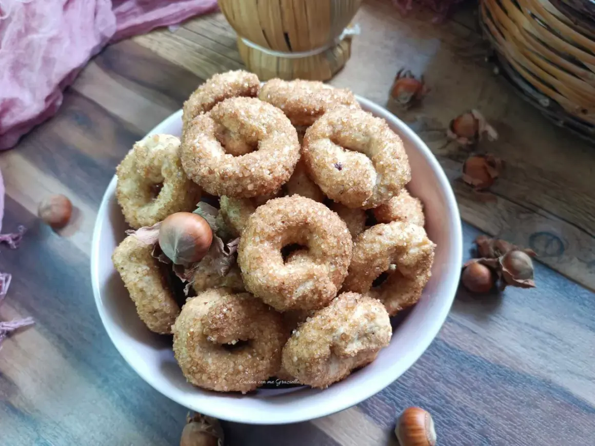 Ciambelline al vino ricetta originale, dorate e croccanti, con nocciole sullo sfondo.