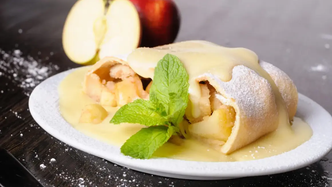 Ein köstlicher Apfelstrudel mit Eiermilch, bestäubt mit Puderzucker und garniert mit Minze. Daneben liegen Äpfel.