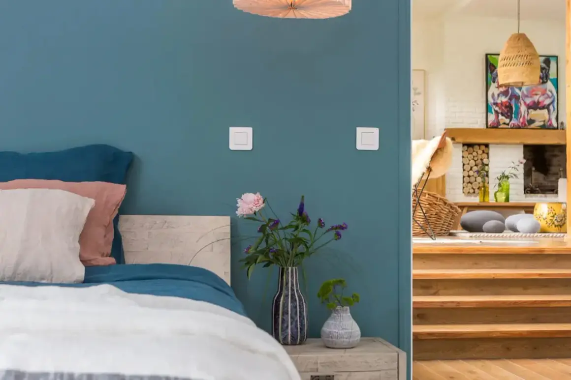 Un lit douillet avec des coussins roses et blancs sur un mur couleur bleu canard. À côté, un escalier en bois mène à un salon cosy.