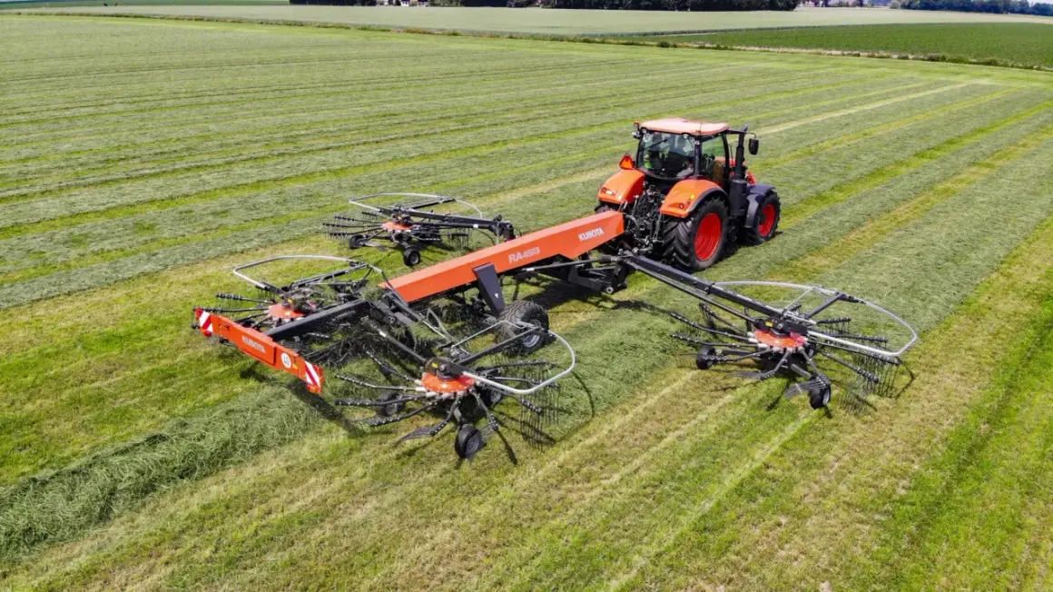 Pomarańczowy traktor zgrabnie manewruje po polu, ciągnąc szeroką zgrabiarkę do traktorka, która sprawnie formuje pokosy zielonki.