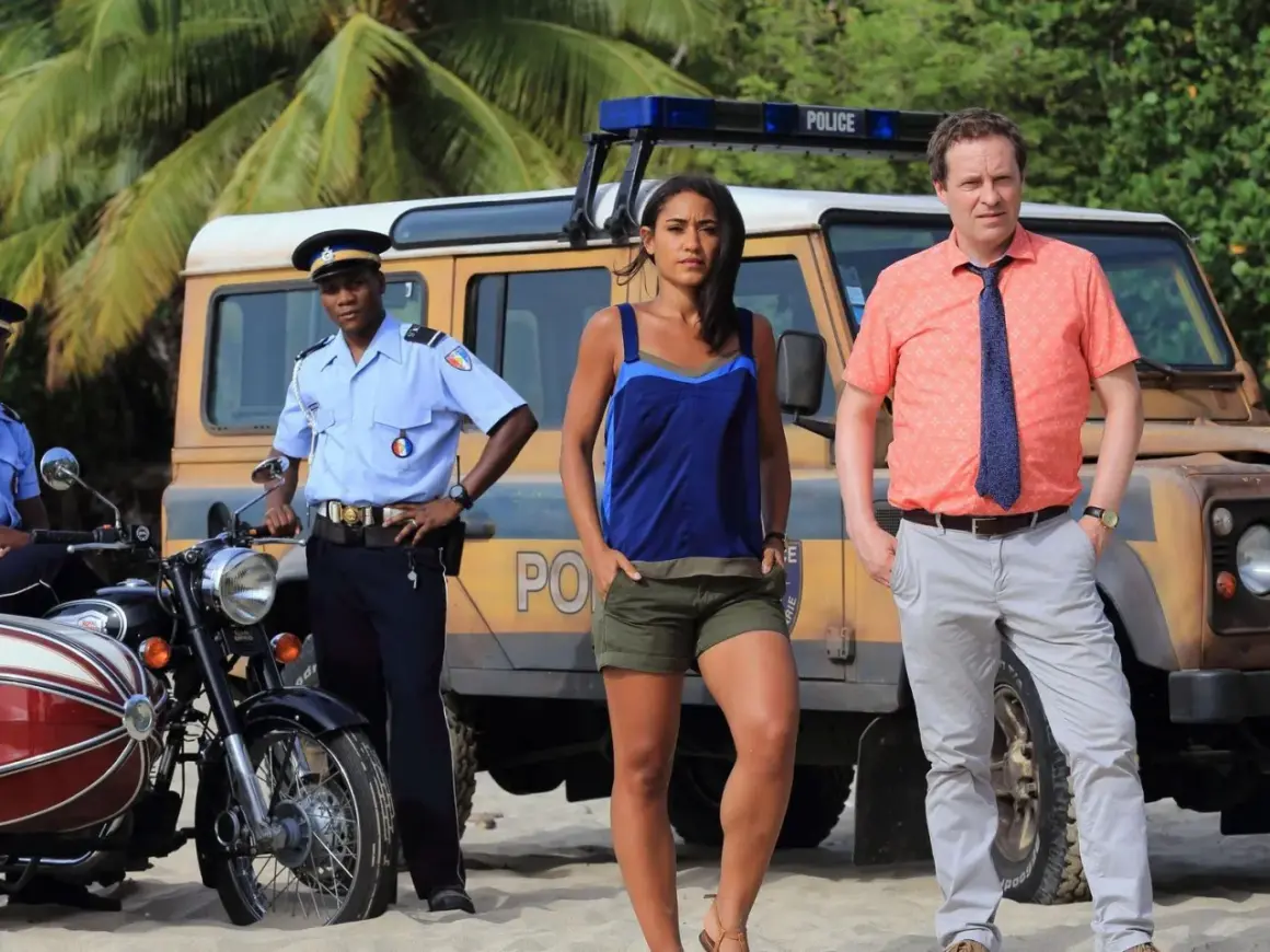 Polizisten und Ermittler am Strand, bereit, "Death in Paradise" online anzusehen. Ein Jeep und ein Motorrad stehen bereit.