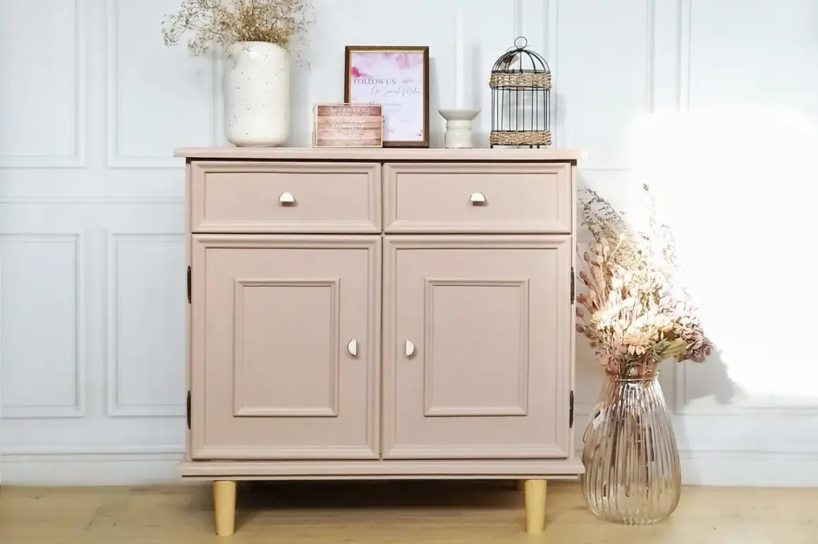 Un meuble en bois, fraîchement peint en rose poudré, est décoré de fleurs séchées et d'une cage à oiseaux.