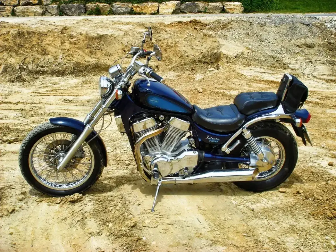 Błękitny motocykl Suzuki Intruder z chromowanymi elementami i czarnym siedzeniem stoi na piaszczystym podłożu.