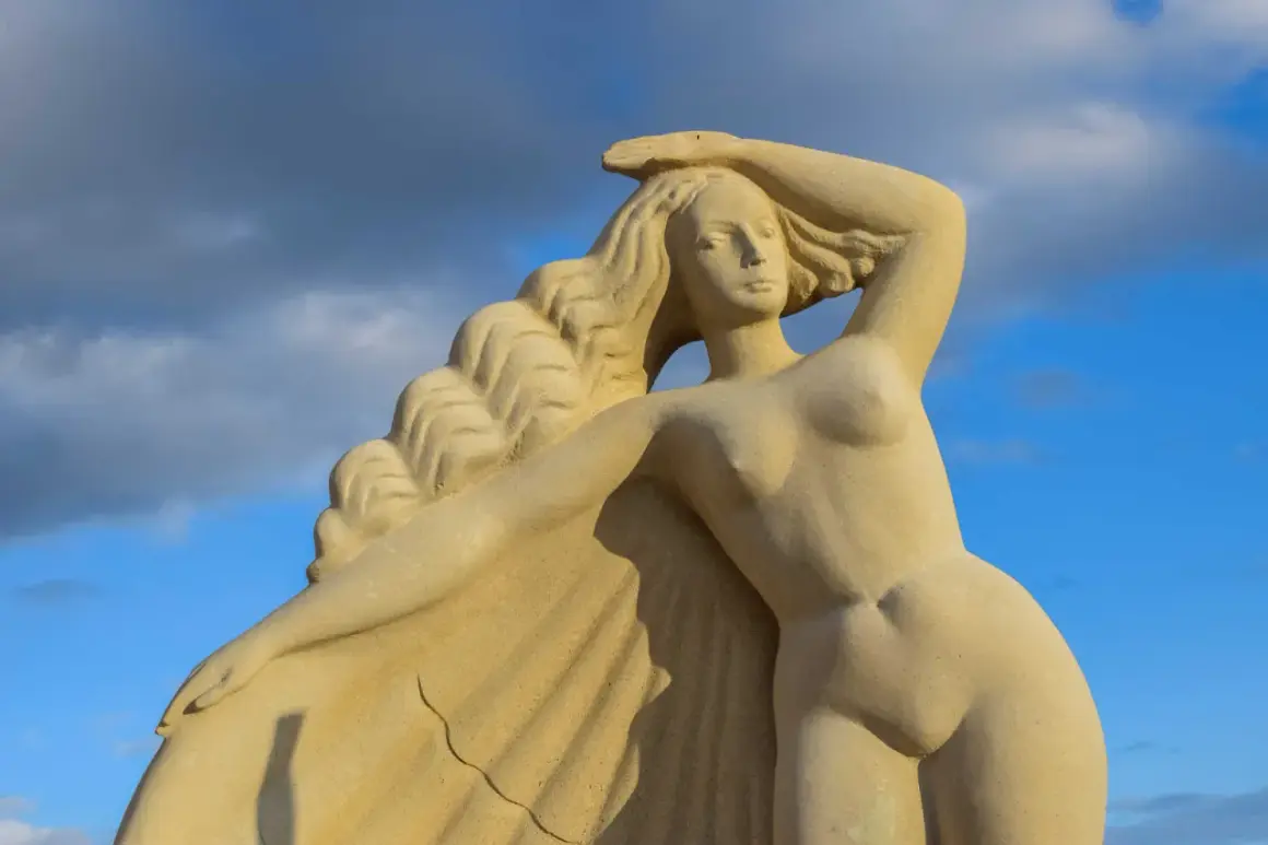 Zandstenen beeld van een **Griekse godin** met golvend haar, tegen een blauwe lucht met wolken.