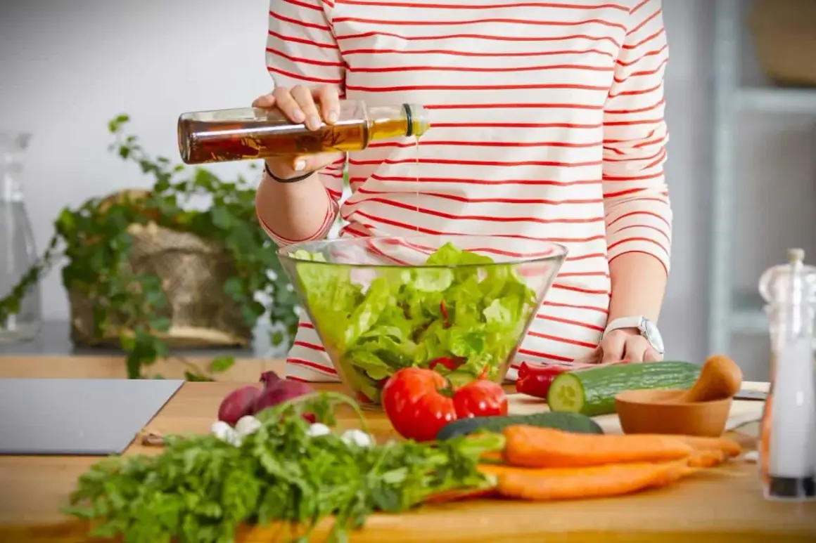 Eine Person bereitet einen Salat zu und träufelt Öl hinein. Frisches Gemüse wie Tomaten, Gurken und Karotten liegen auf dem Tisch. Ein Hauch von weisswein essig rundet das Gericht ab.