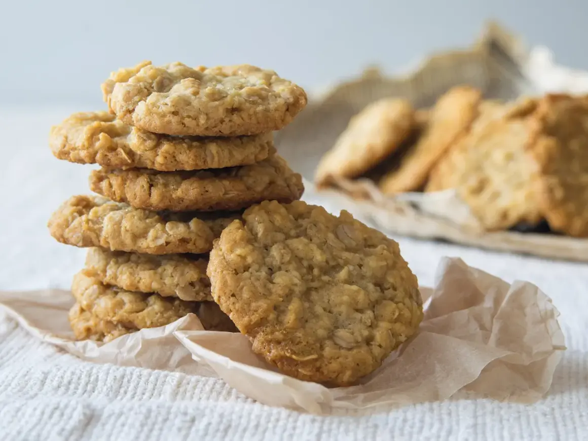 Ein Stapel goldbrauner Haferflocken-Bananen-Cookies auf einem weißen Tuch, daneben weitere Kekse.
