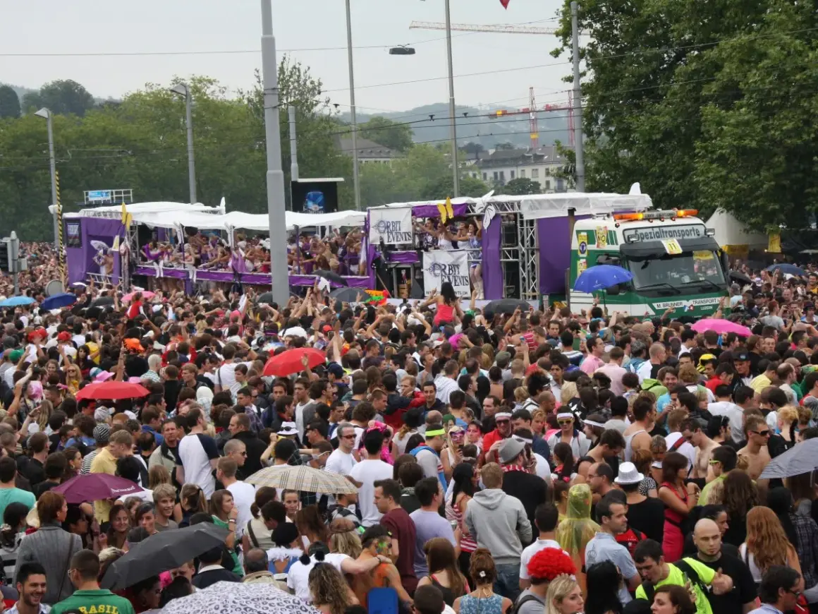 Folla festante a Zurigo per una street parade. Persone danzano e salutano dai carri colorati, un camion speciale trasporta la musica.