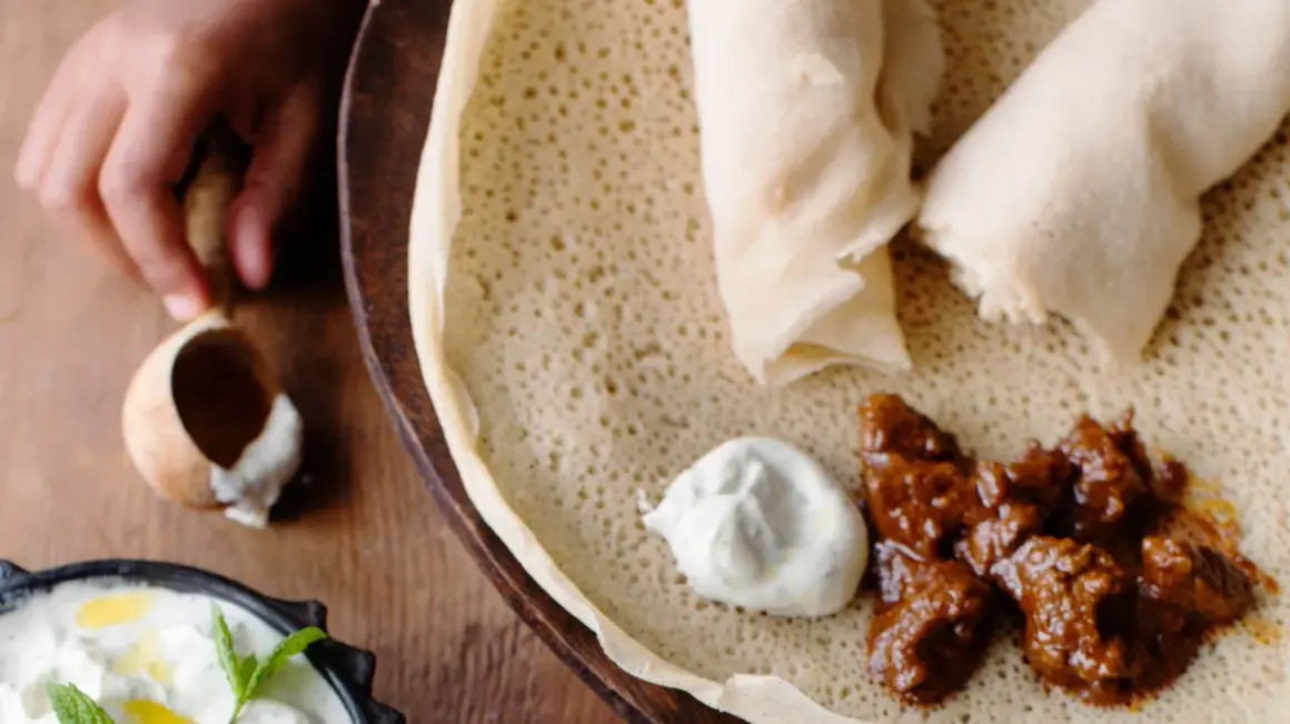 En hand tar en bit av en injera, en etiopisk rätt med kött och sås. Ett perfekt injera recept för en smakrik måltid.