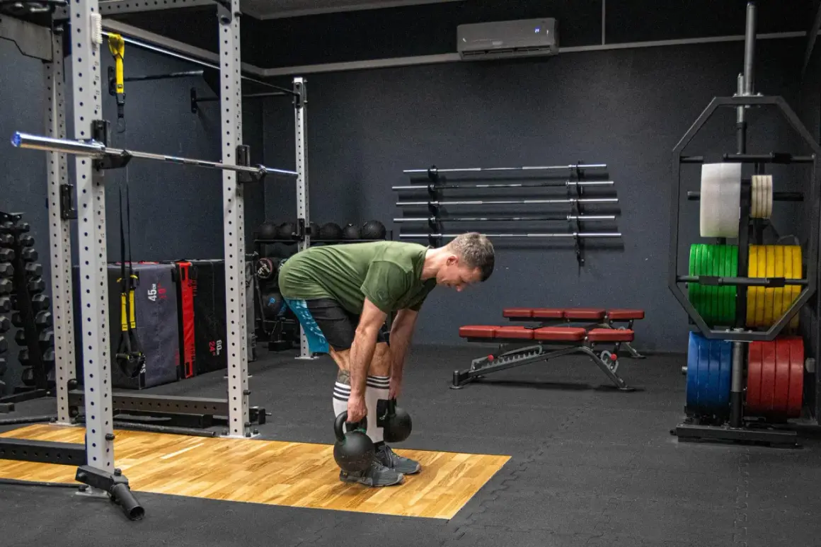 Mężczyzna wykonuje martwy ciąg rumuński z kettlebellami, ćwicząc mięśnie tylnej taśmy. W tle widać sprzęt do treningu siłowego, w tym sztangi i obciążenia.