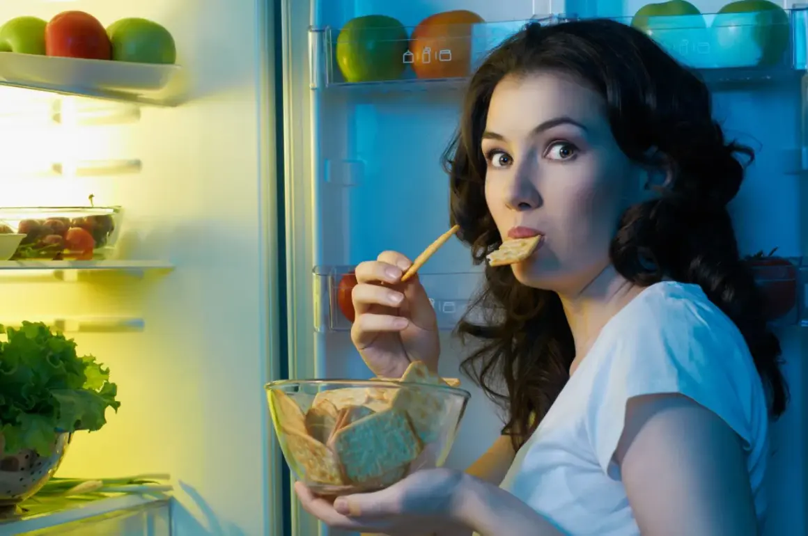 Une femme, prise par une **sensation de faim**, grignote des biscuits devant un réfrigérateur ouvert, rempli de fruits et légumes.