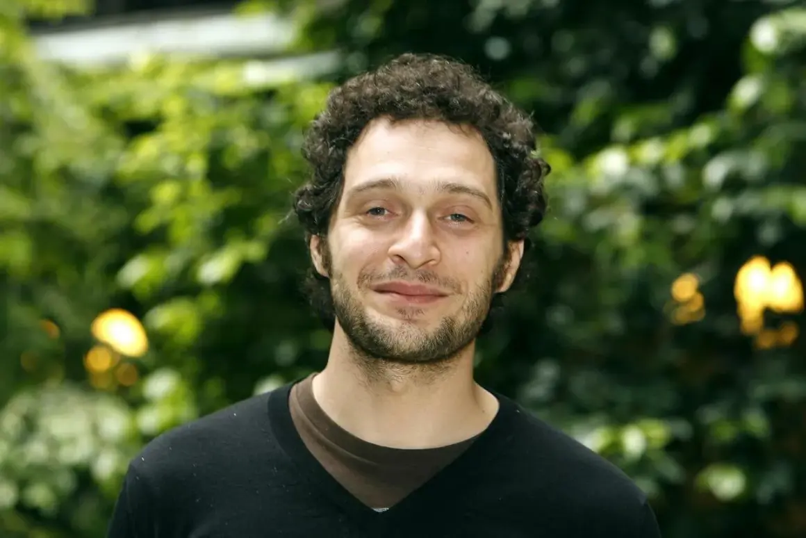 Claudio Santamaria giovane, con capelli ricci e barba, sorride con uno sfondo verde sfocato.