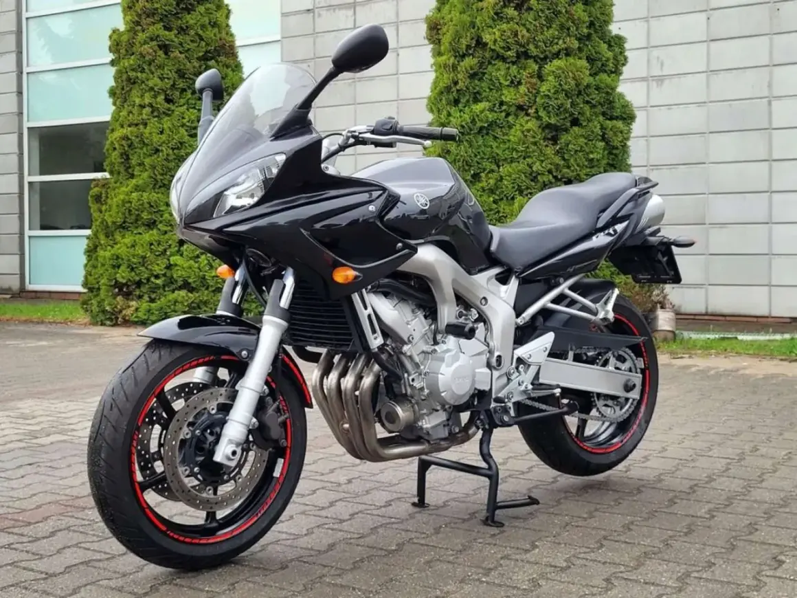 Czarny motocykl Yamaha FZ6, gotowy do jazdy, z czerwonymi akcentami na felgach, stoi przed budynkiem z zielonymi drzewami.