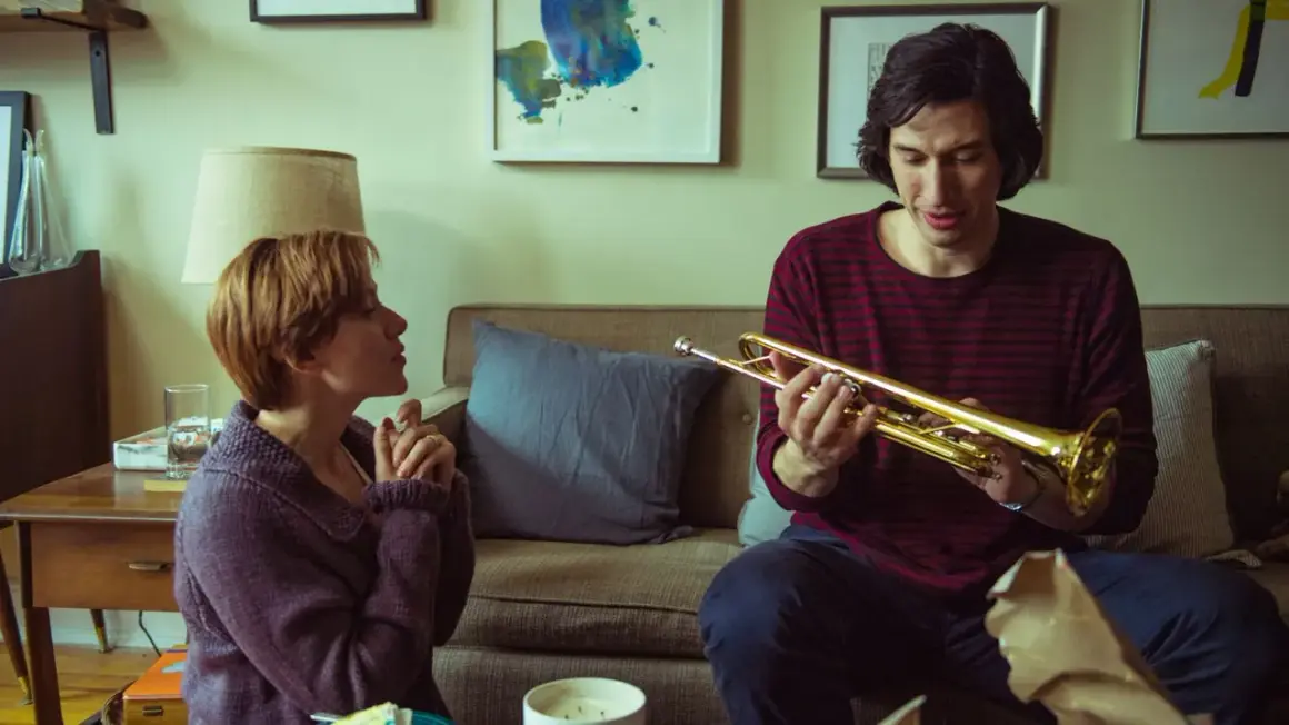 Adam Driver håller i en trumpet i en scen från en av hans filmer. Bredvid honom sitter en kvinna och tittar på.