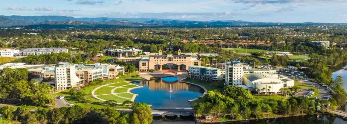Weite Luftaufnahme des Campus der Bond University mit einem zentralen See, gepflegten Grünflächen und modernen Gebäuden, umgeben von Bäumen und Hügeln.