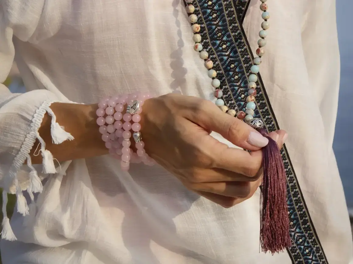 Mano con varios tipos de japa mala: uno de cuarzo rosa en la muñeca y otro largo con cuentas de colores y borla.