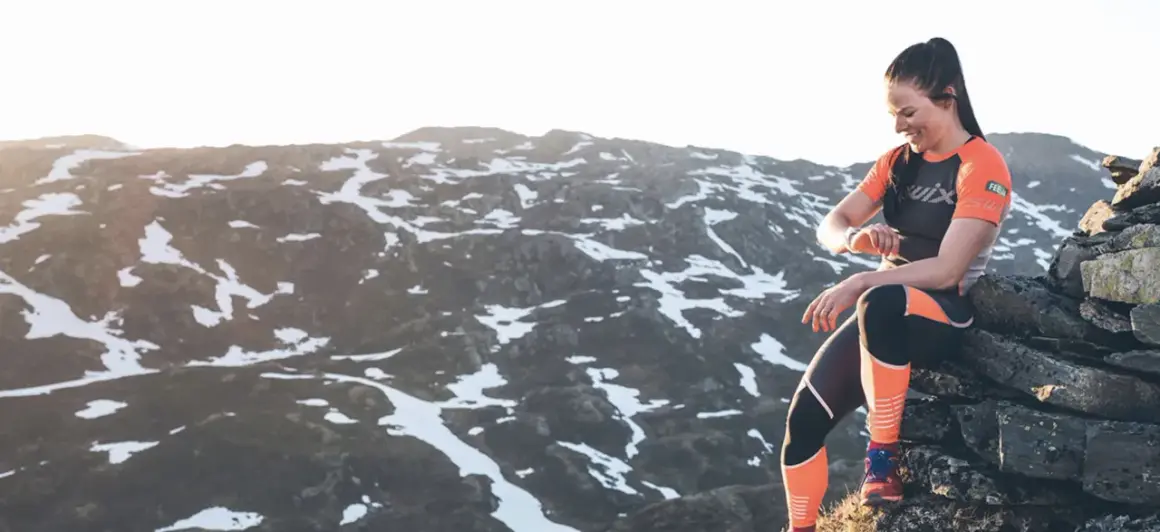 Une femme en tenue de sport s'assoit sur des rochers, regardant sa montre, prête à trouver un trail dans un paysage montagneux enneigé.