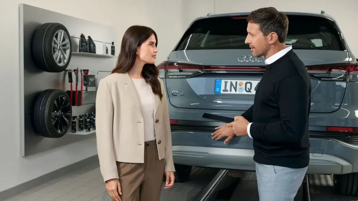 Klientka rozmawia z pracownikiem serwisu Audi o akcjach serwisowych. Na ścianie widoczne opony i akcesoria.