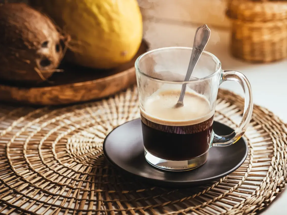 Tazza di caffè fumante su sottobicchiere intrecciato. Sullo sfondo, cocco e melone. Un caffè lungo ha più caffeina per iniziare la giornata.