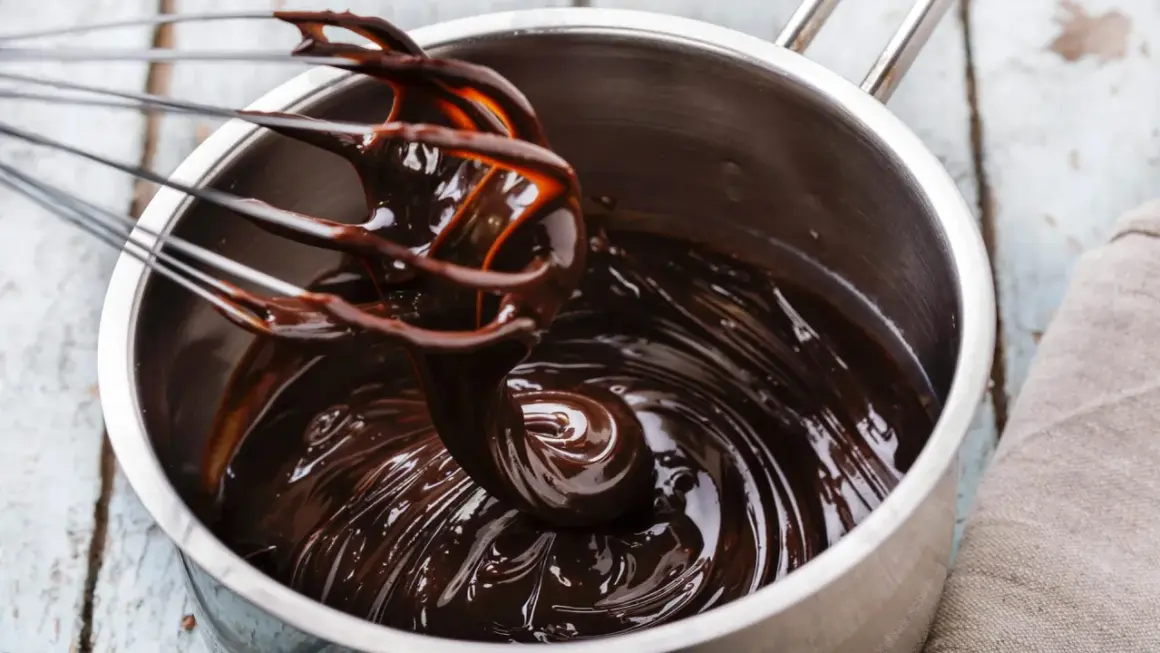 Ein Schneebesen rührt glänzende, dunkle schoko ganache in einem Topf.