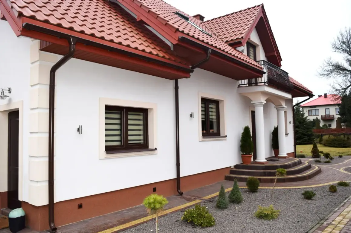 Biały dom z brązowymi rynnami i dachówką. Przed domem ozdobne krzewy i ścieżka z cokolików.