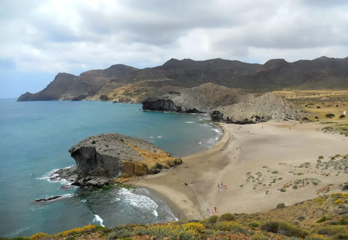 Paisaje de las mejores calas de Cabo de Gata, con formaciones rocosas, arena dorada y mar turquesa bajo un cielo nublado.