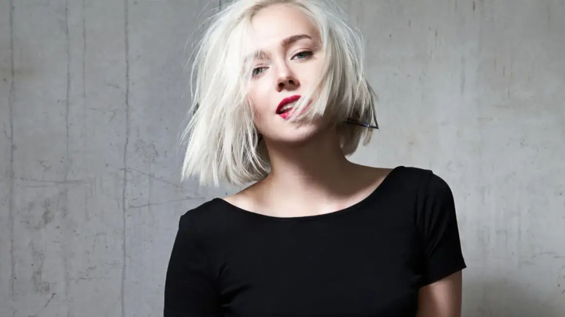 Blonde Frau mit einem geraden Bob und rotem Lippenstift, die provokativ in die Kamera blickt.