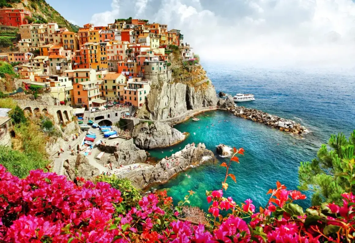 Färgstarka hus klättrar uppför klipporna vid den italienska kusten. Klart turkost vatten och rosa blommor ramar in vyn.