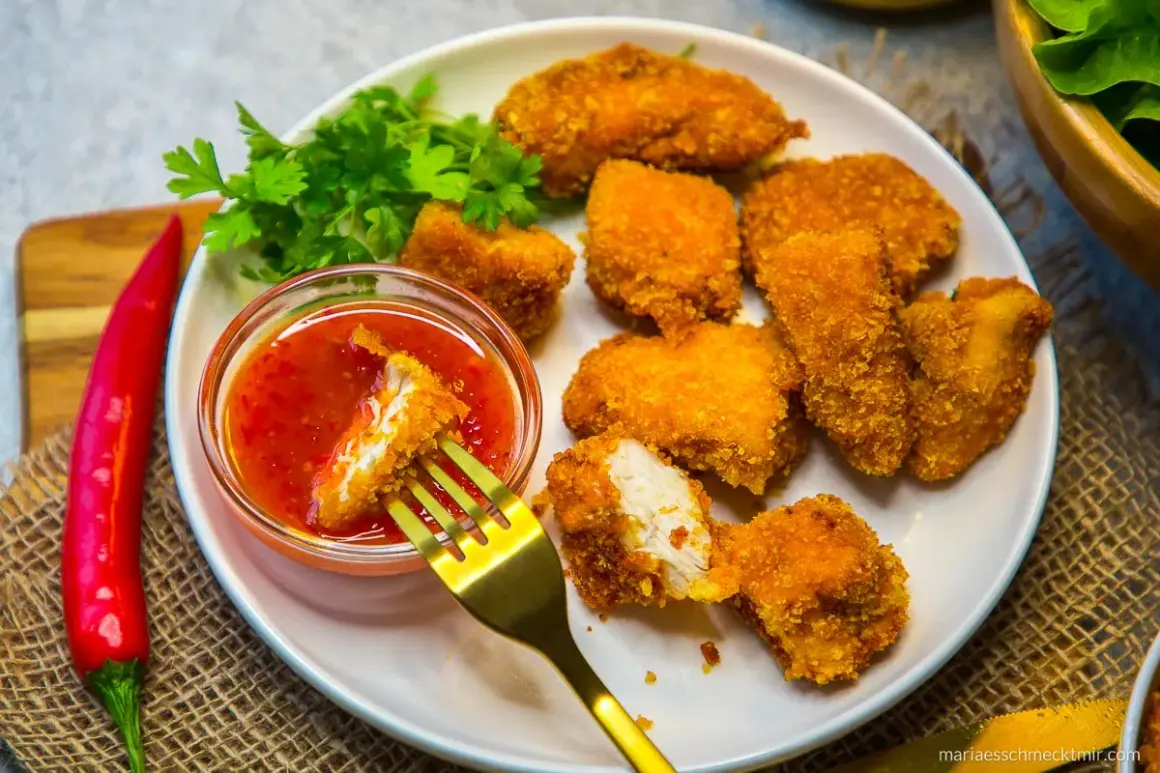 Knusprige Hähnchen-Nuggets, die man einfach selber machen kann, mit süß-saurer Soße und frischer Petersilie. Ein roter Chili liegt daneben.