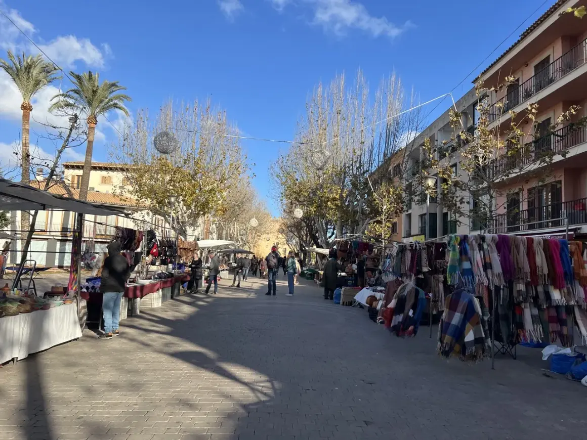 Livlig marknad i Alcudia med stånd som säljer kläder och accessoarer. Palmträd och byggnader kantas av gatan.