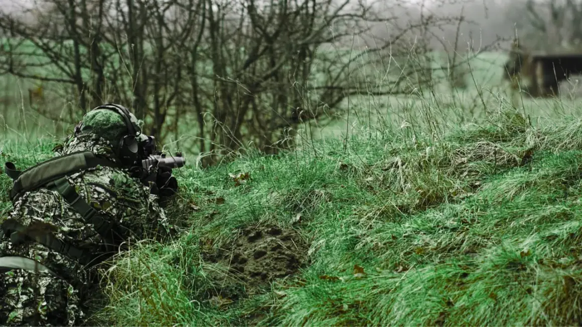 Jugador de airsoft que dispara desde la hierba alta, camuflado y con equipo táctico.